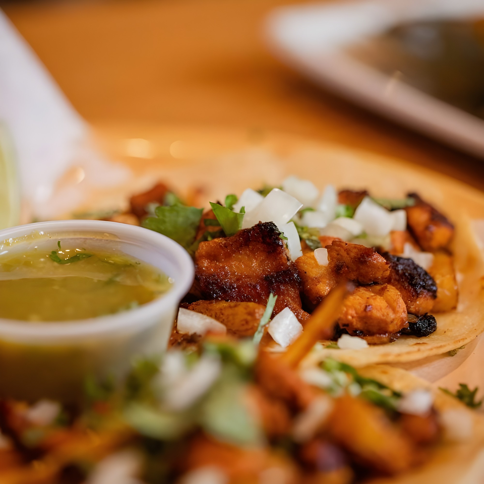 Close-up of three tacos with grilled meat, diced onions, cilantro, and a side of green salsa.
