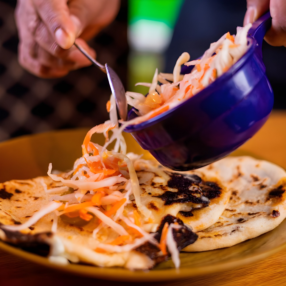 A hand spoons curtido from a blue bowl onto two pupusas on a brown plate.